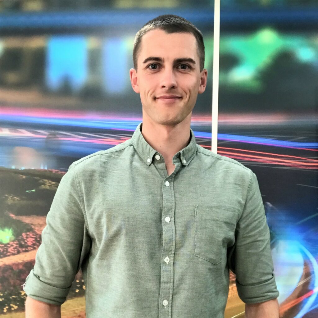 A headshot photo of Bradley Goodsell in front of a vibrantly coloured branded SYSTRA office wall. He is smiling at the camera and is wearing a a pale green shirt.