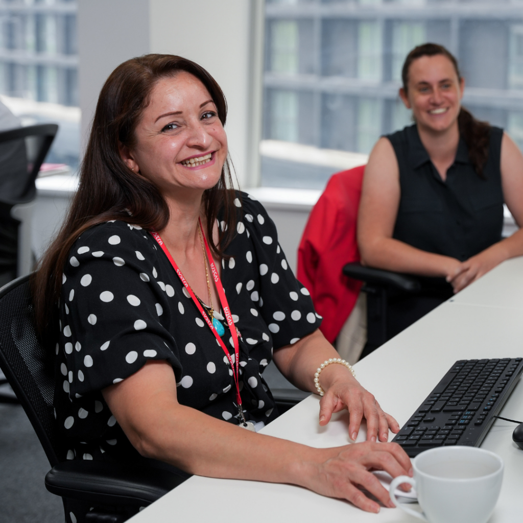 A photo of a female SYSTRA colleague sat at her desk. She is wearing a black and white spotty top, her red lanyard and is smiling at the camera. In the background out of focus is another female SYSTRA colleague looking away, smiling.