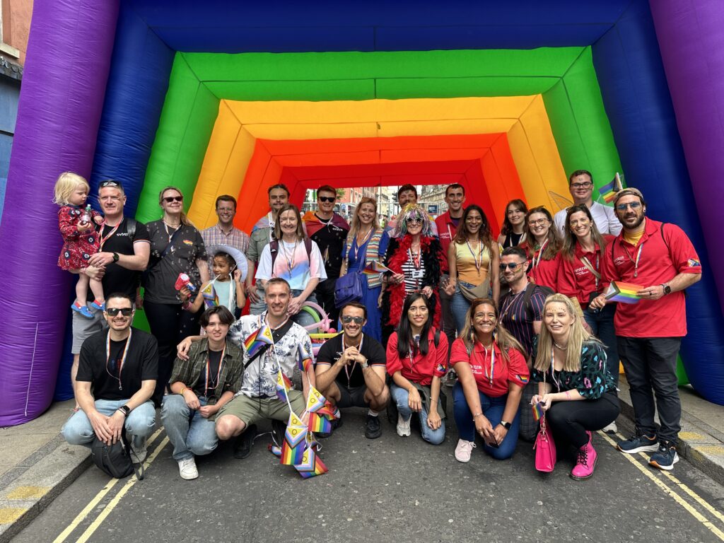 A big group of SYSTRA colleagues, family and friends pictured in front of a multicoloured inflatable at Leeds Pride 2024. They are all wearing bright colours and smiling at the camera.