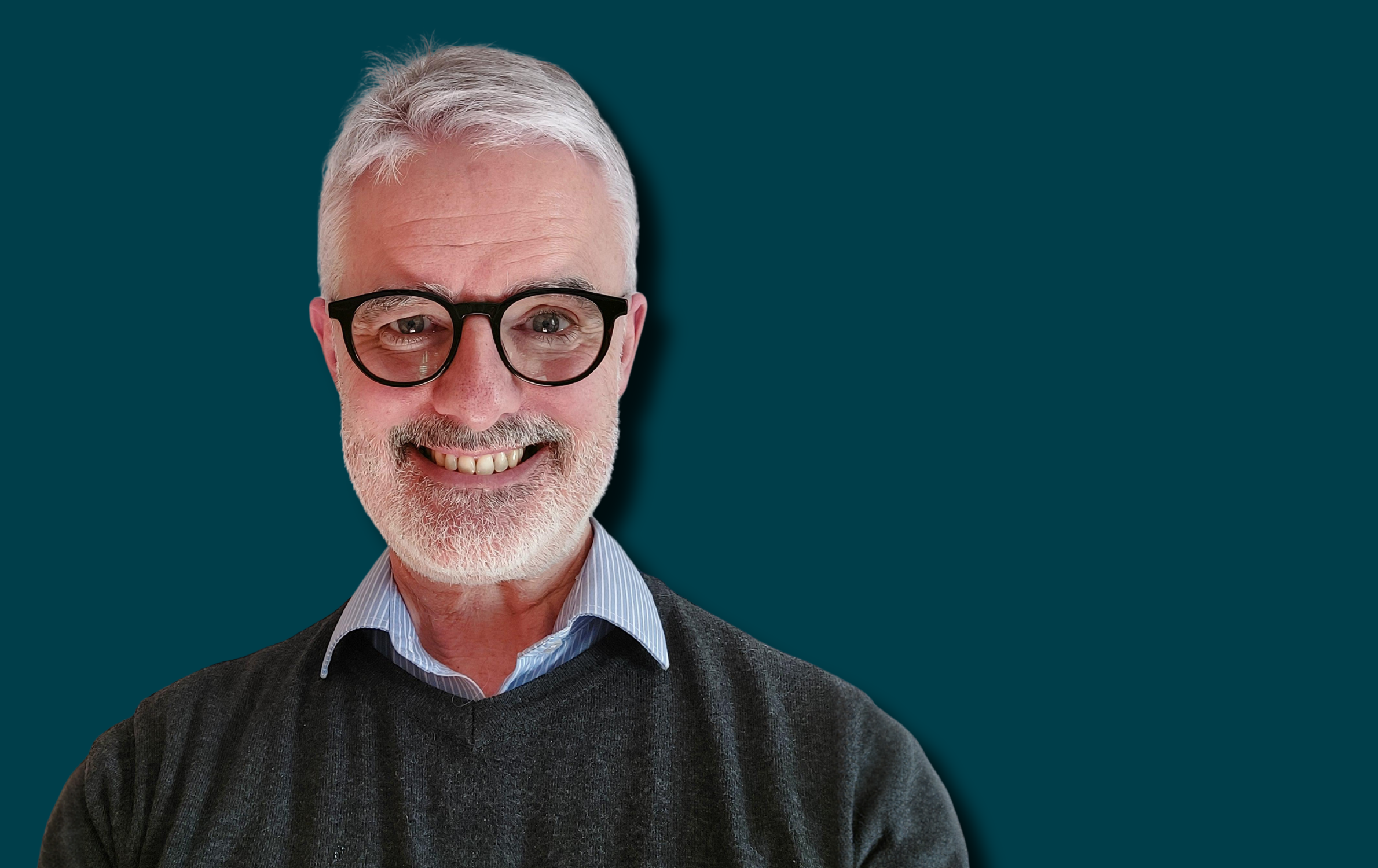 A headshot photo of Ian Byrne who is wearing a dark grey jumper, a blue shirt, glasses, and is smiling at the camera.