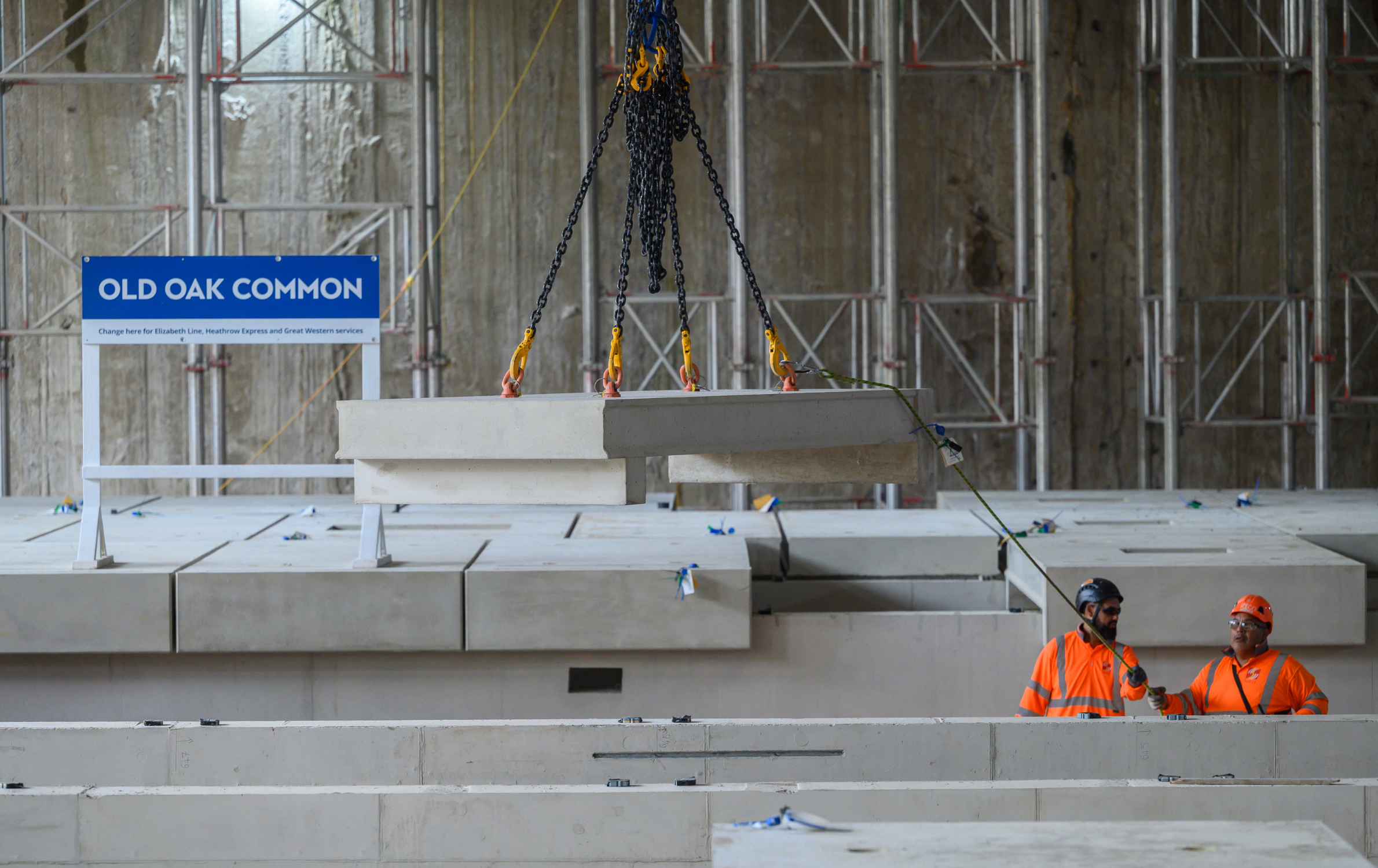 A photograph of two workers at the HS2 Old Oak Common station construction site where the first platforms are being installed. They are wearing hard hats and bright orange PPE, and are stood below the platform level where the tracks run. On the platform there is a bright blue sign that says Old Oak Common.