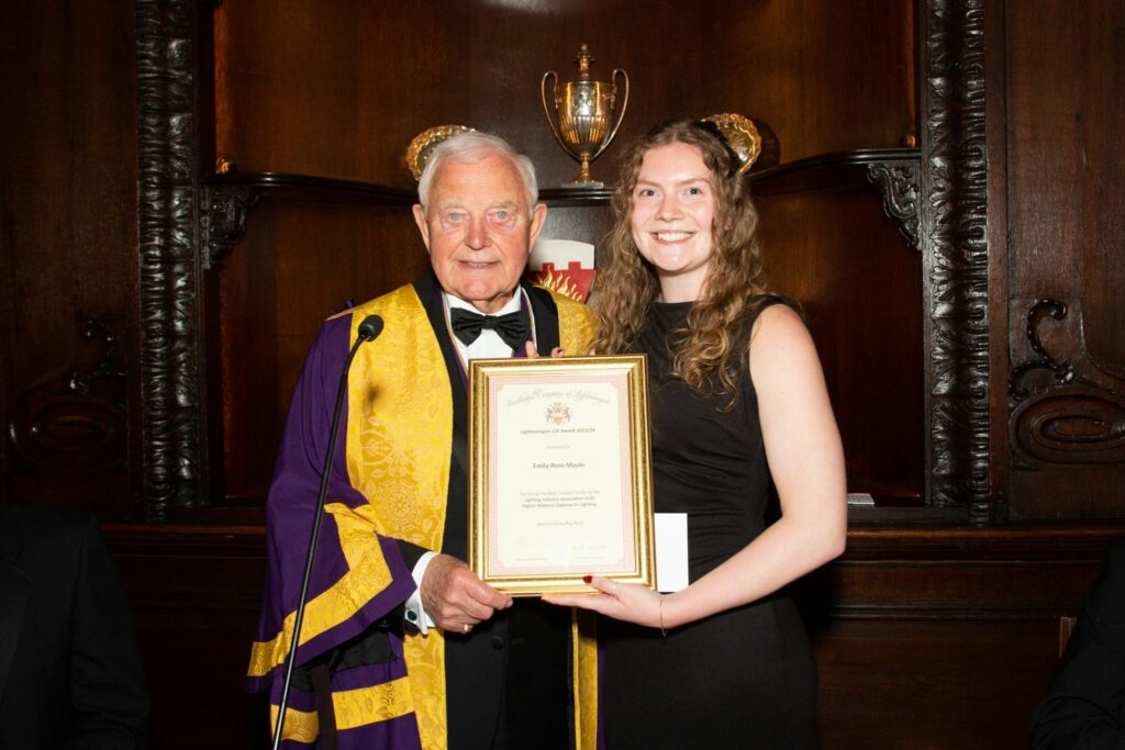 Emily Rose-Maule is pictured receiving her LIA Advanced Exterior Lighting Design Certificate Outstanding Student of the Year award. She is standing next to a man in official clothing and is holding her award. She is smiling at the camera.