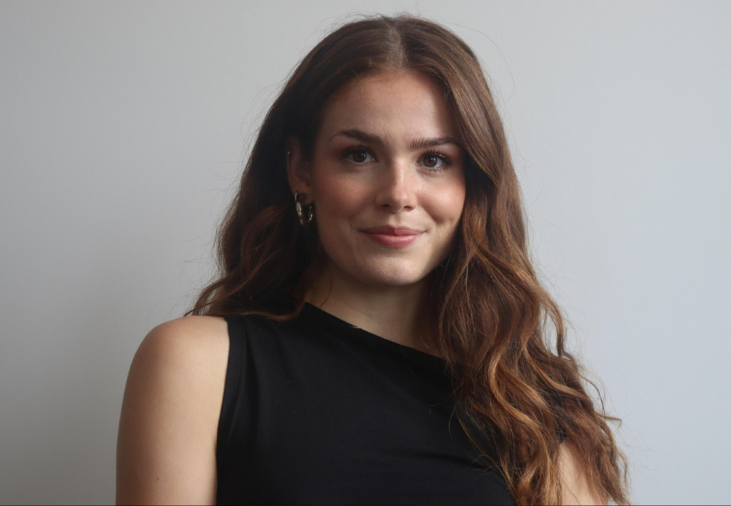 A professional headshot photo of Riona Ainge (Sustainability Lead at SYSTRA). Riona is wearing a black top and is looking at the camera smiling. The background is grey.