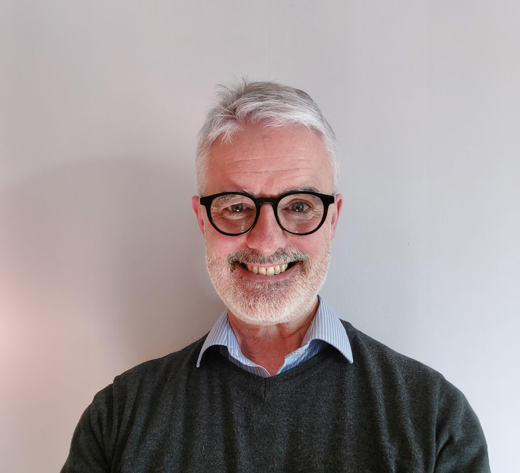 Headshot photo of Ian Byrne. He is wearing a blue shirt and a black jumper, and is smiling at the camera.