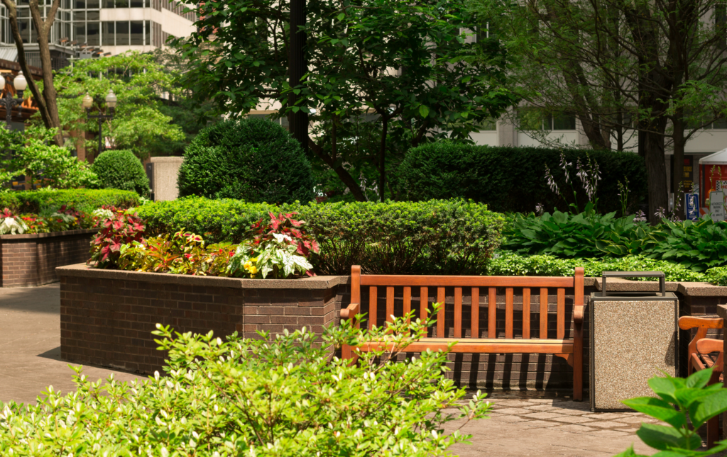 A wooden bench in a small pocket park in a city. The bench is in front of a brick wall and is surrounded by bushes and flowers.
