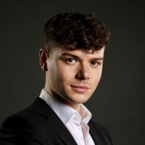A headshot photo of Joe Thompson, PlanBEE Rail apprentice. Joe is wearing a white shirt and a black suit jacket, and is looking at the camera. The background is dark.