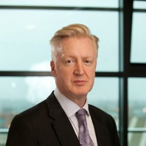 A headshot photo of Paul Chelin who is standing in front of a large window, looking at the camera. He is wearing a shirt and tie and a black suit jacket.