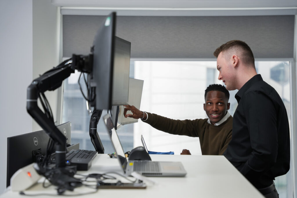 A photo of 2 SYSTRA colleagues talking in a SYSTRA office. They are both facing a computer screen, and one of them is pointing at it and smiling.