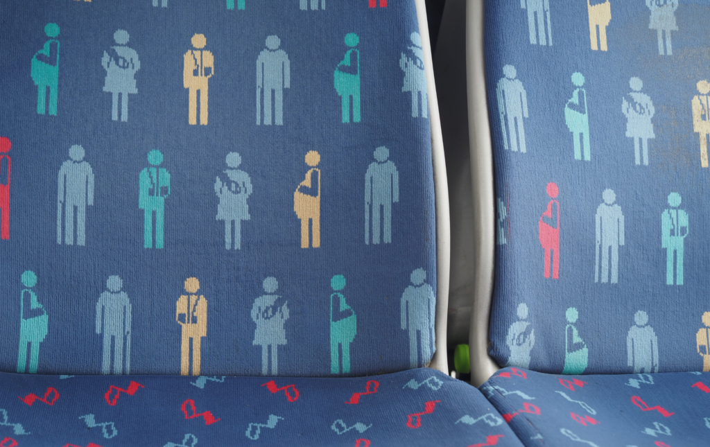 Zoomed in photo on priority seat on train. The seat is blue and has a colourful pattern on it that shows various examples of people that require usage of the seat for accessibility reasons - for example a pregnant person, person with a sling, person with a walking stick, person carrying a baby.