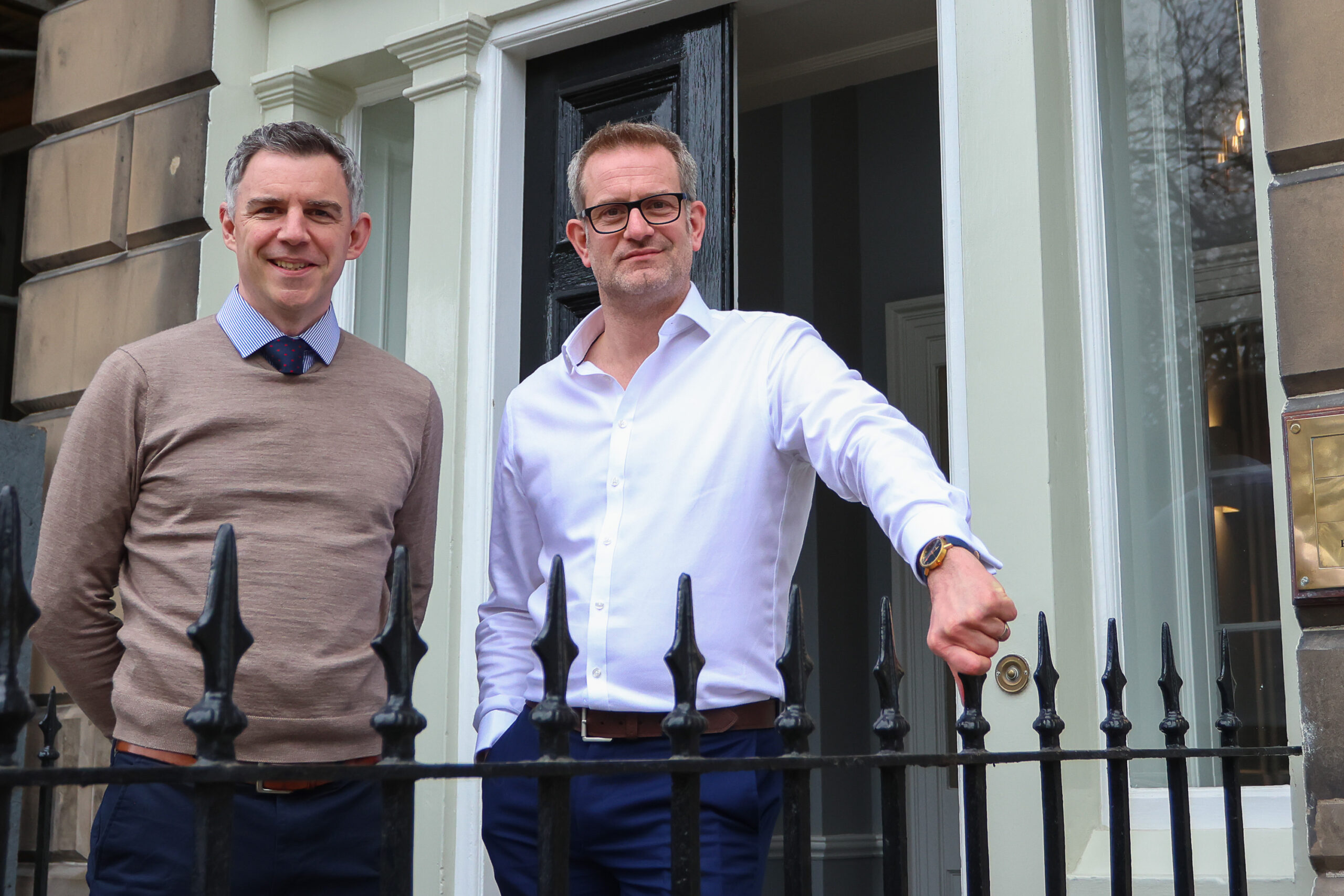 Photo of Keith McGillivray standing outside the entrance to the SYSTRA Edinburgh office next to a colleague. He is wearing  white shirt and black suit trousers, and has his hand on a black metal fence. His colleague is wearing black suit trousers and a brown jumper. They are both smiling at the camera.