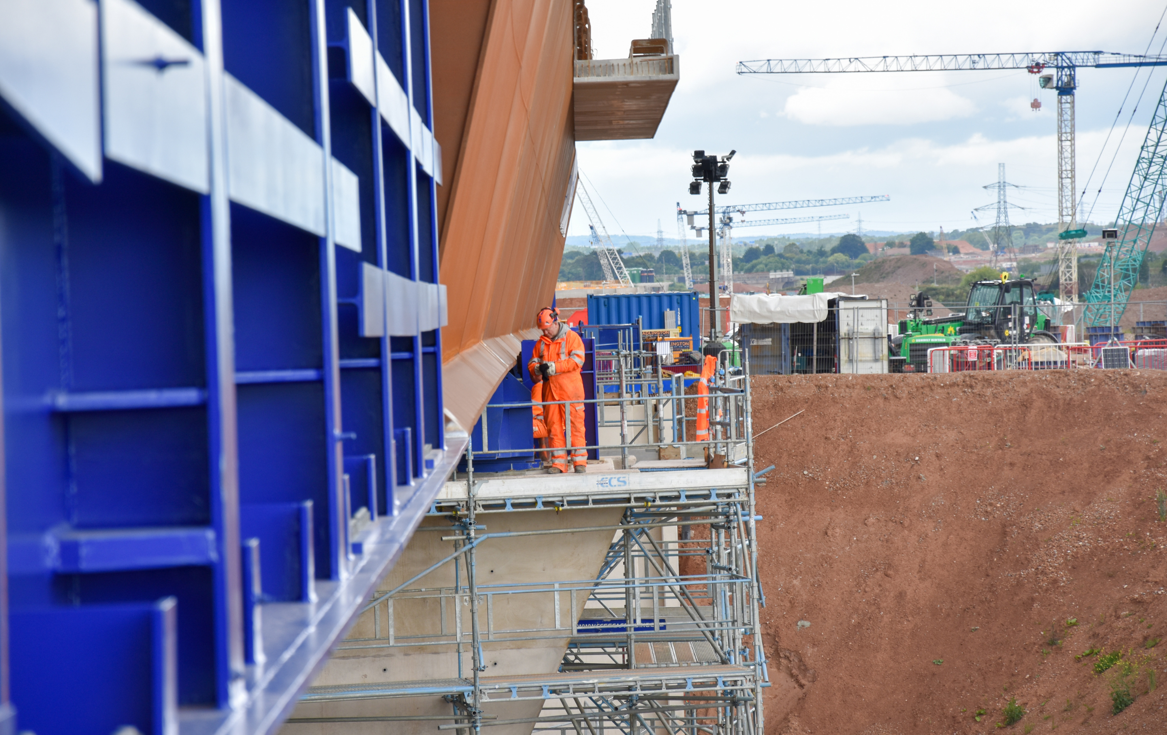 View along the side of the M6 South viaduct deck as it's being installed on the HS2 project. A construction worker wearing PPE is pictured on some scaffolding next to the large structure. The construction site is in the background.