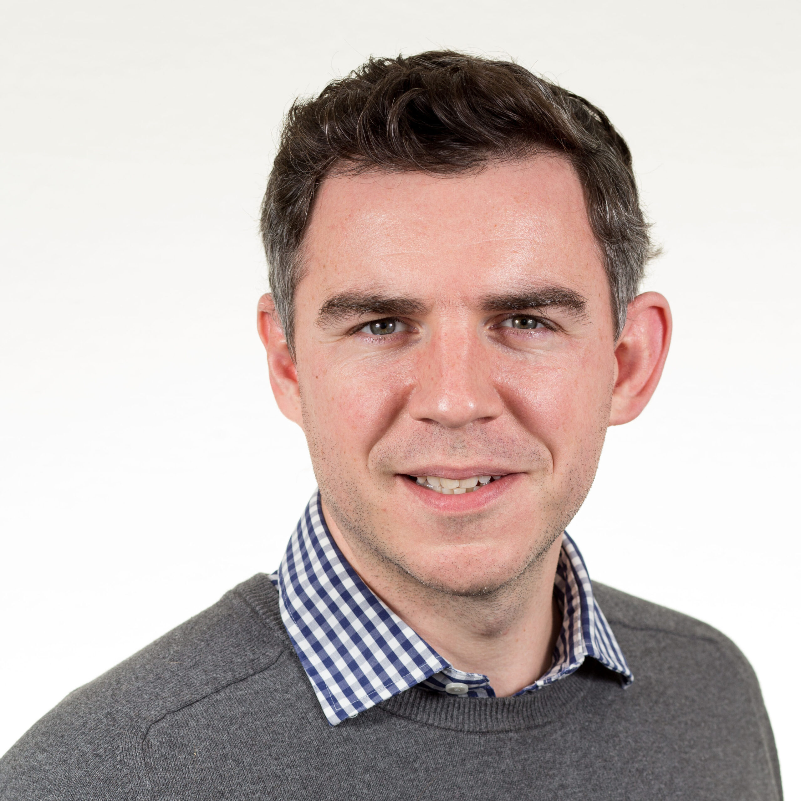 A headshot image of Malcolm Calvert. Malcolm is wearing a grey jumper and is smiling at the camera. The backdrop is white.
